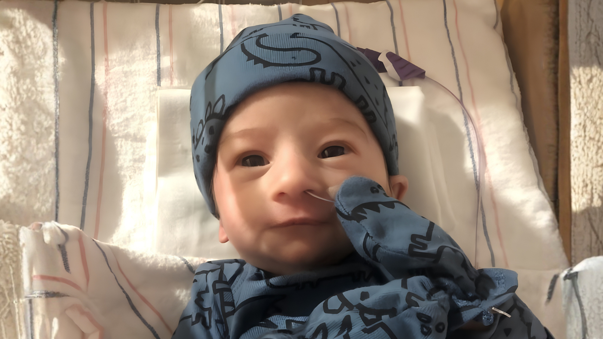 Newborn baby wearing a blue hat and outfit, lying in a hospital bassinet with a nasal feeding tube.