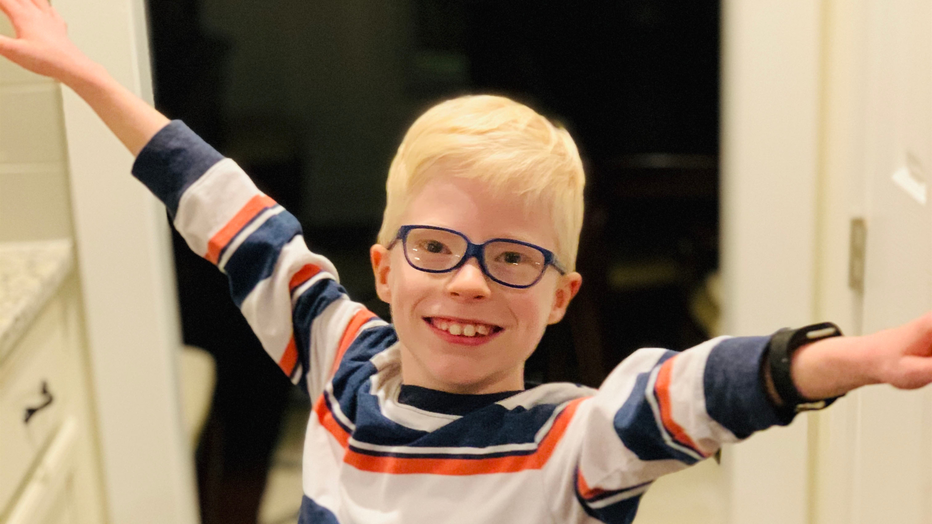 Smiling young boy with glasses holding his arms out wide.