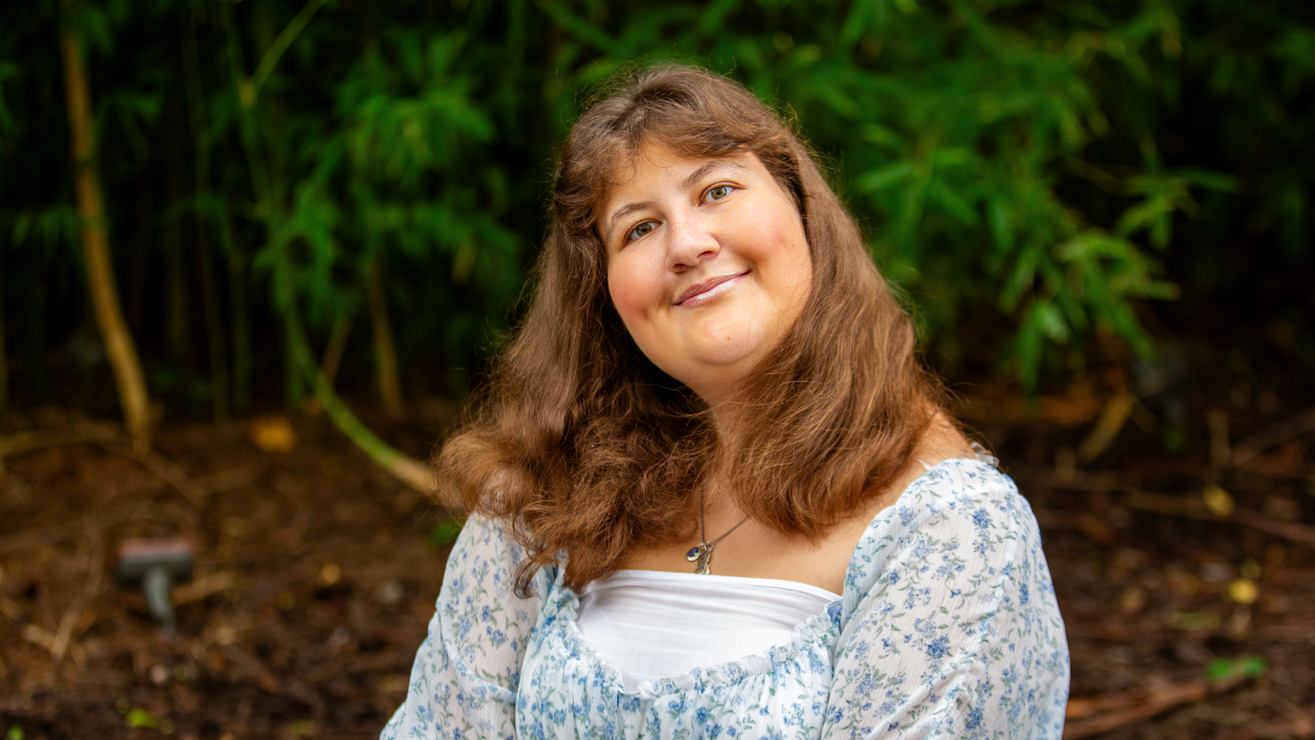 Smiling young woman with long brown hair posing outdoors in front of green foliage.