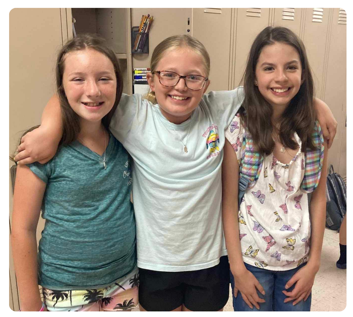Three girls smiling together with their arms around each other indoors.