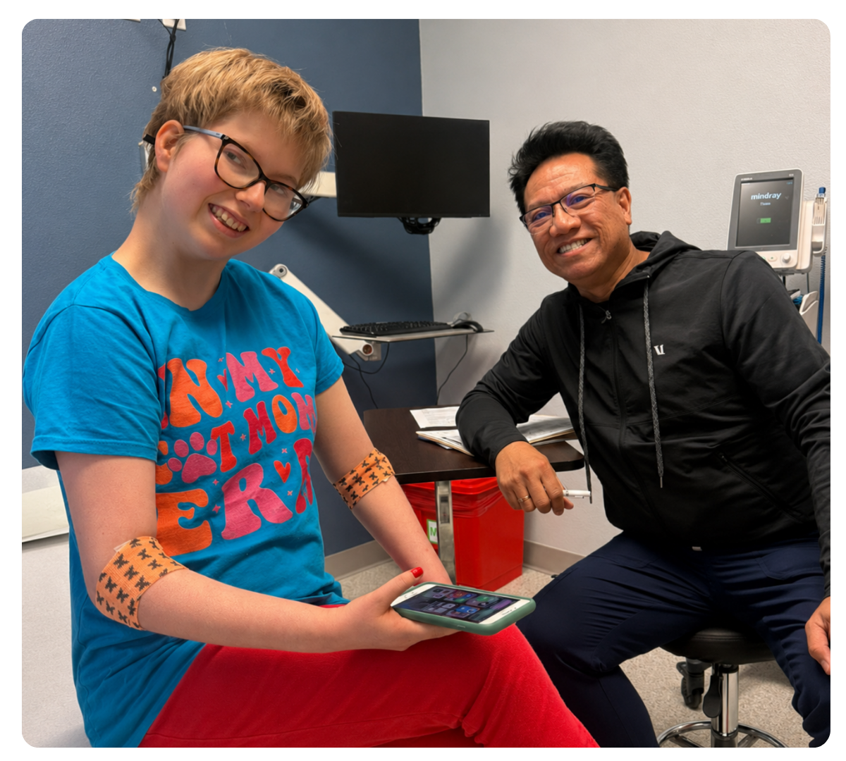 Young woman wearing glasses sits beside a smiling healthcare professional in an exam room, with monitoring patches on her arms.