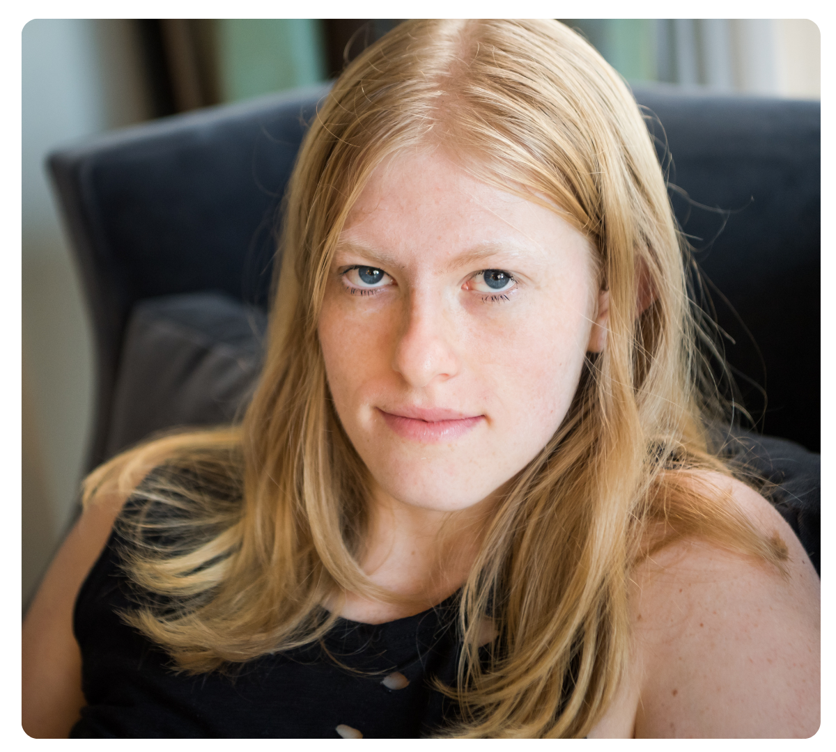 Young woman with long blonde hair looks at the camera while seated indoors.
