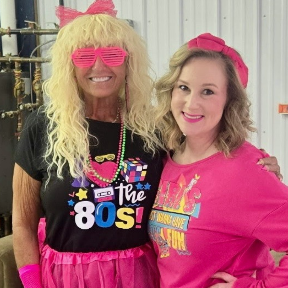 Two women smiling together in colorful 1980s-themed outfits at a fundraising event.