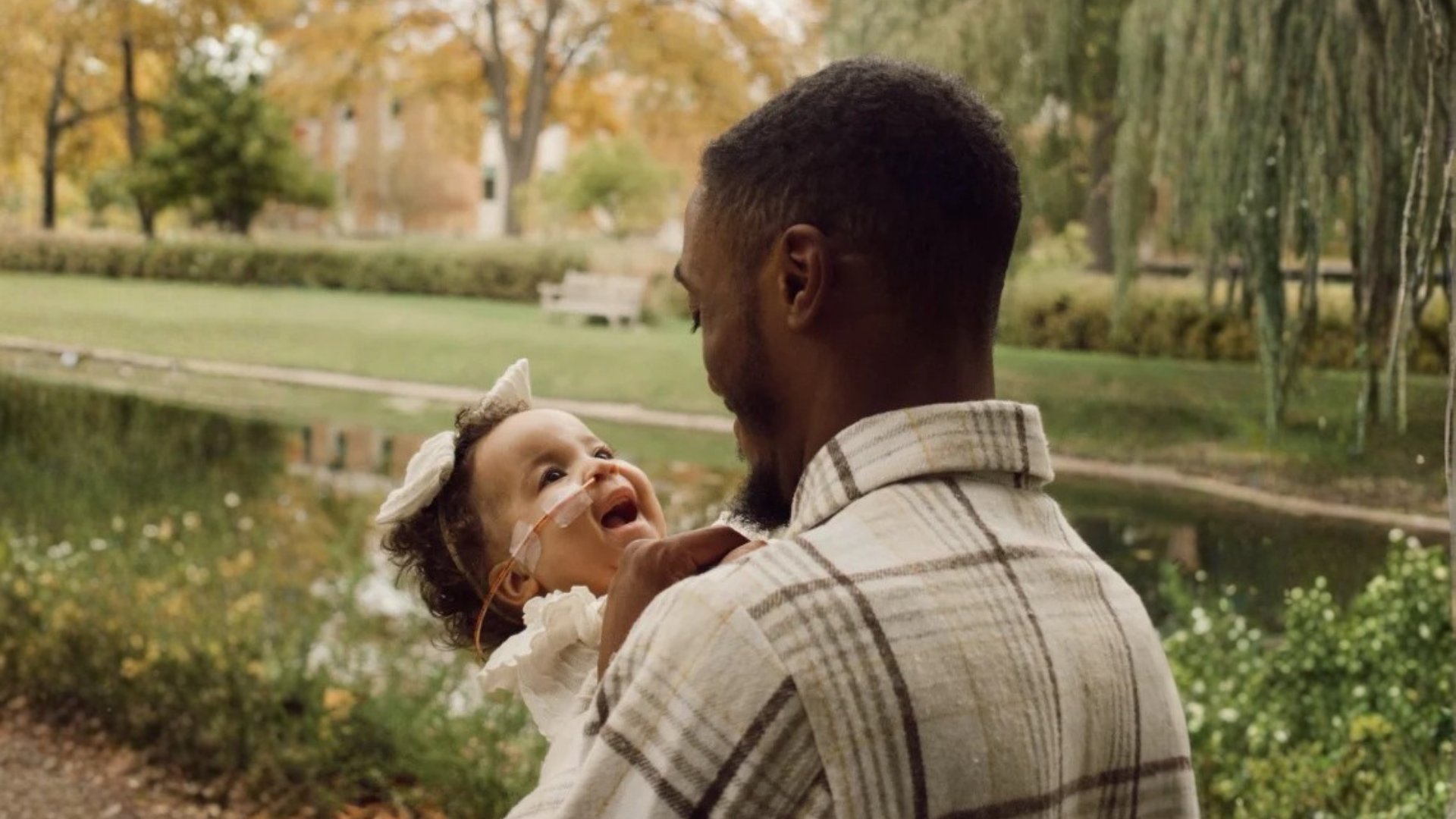Father holding his smiling baby, who wears a nasal feeding tube, beside a pond in a park.