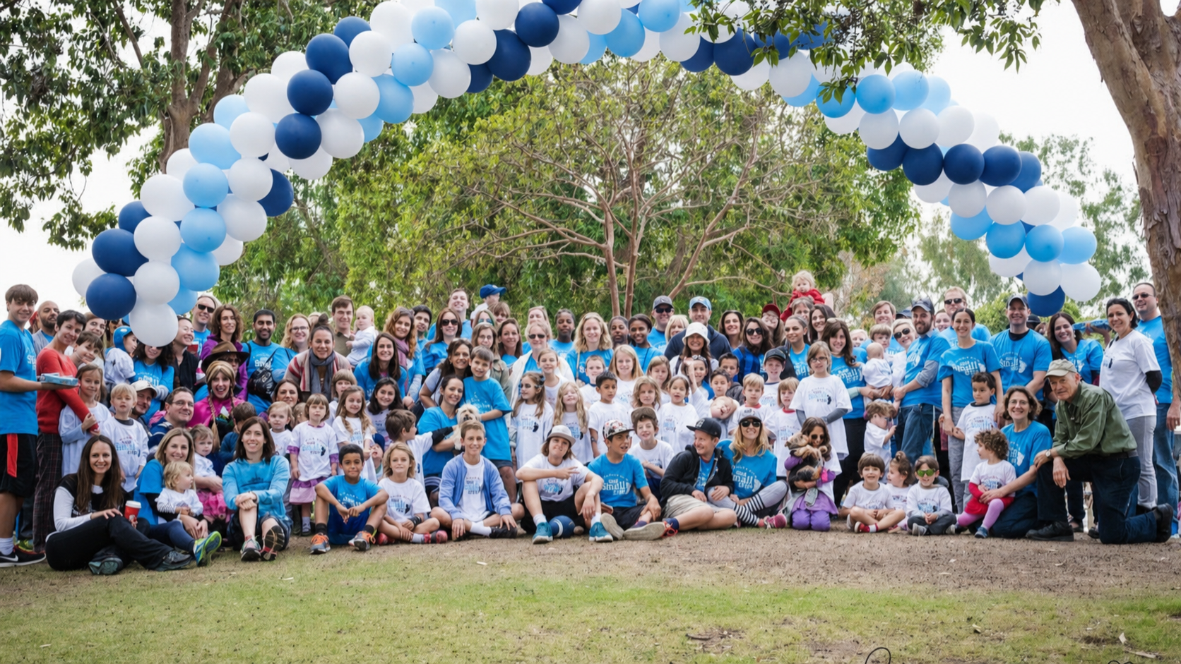 A large group of children and adults pose beneath a blue-and-white balloon arch at an outdoor One Small Step walk.