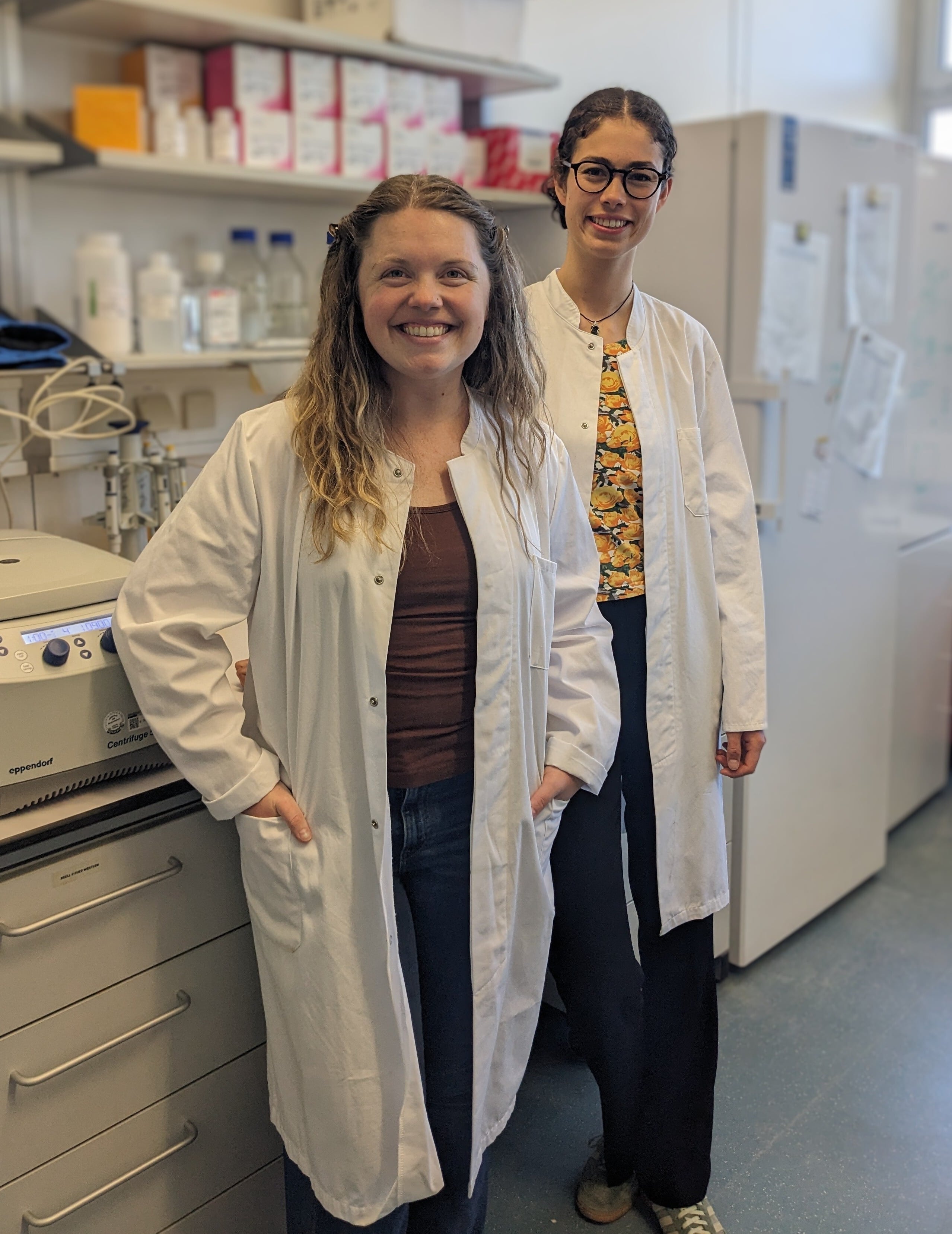 Two researchers wearing white lab coats smiling together in a laboratory.