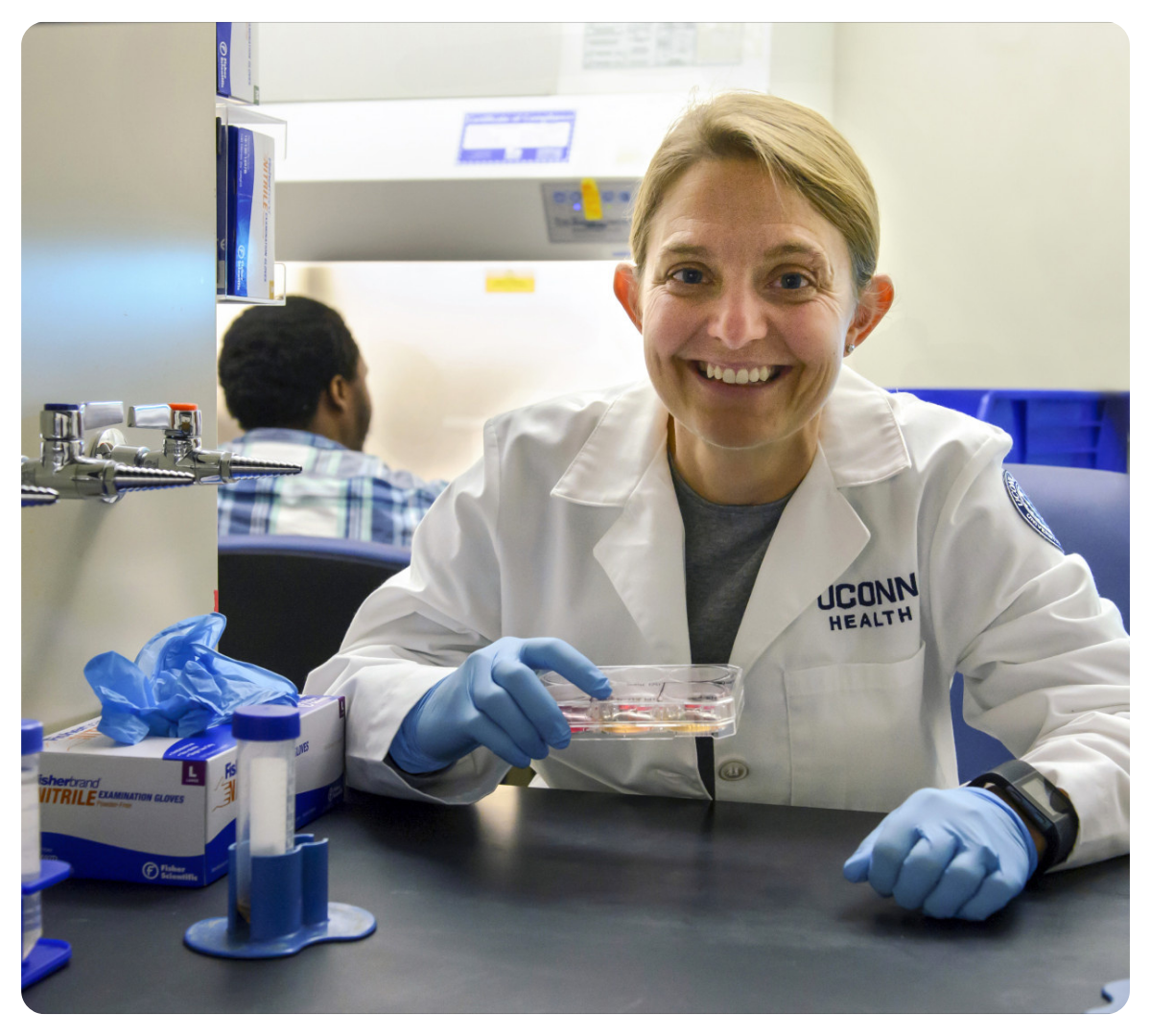 Researcher in a lab coat smiles while holding a cell culture plate in a laboratory.