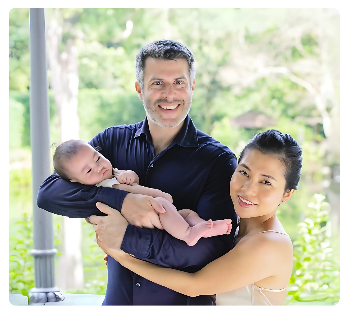 A man and woman smiling as they hold a newborn baby outdoors.