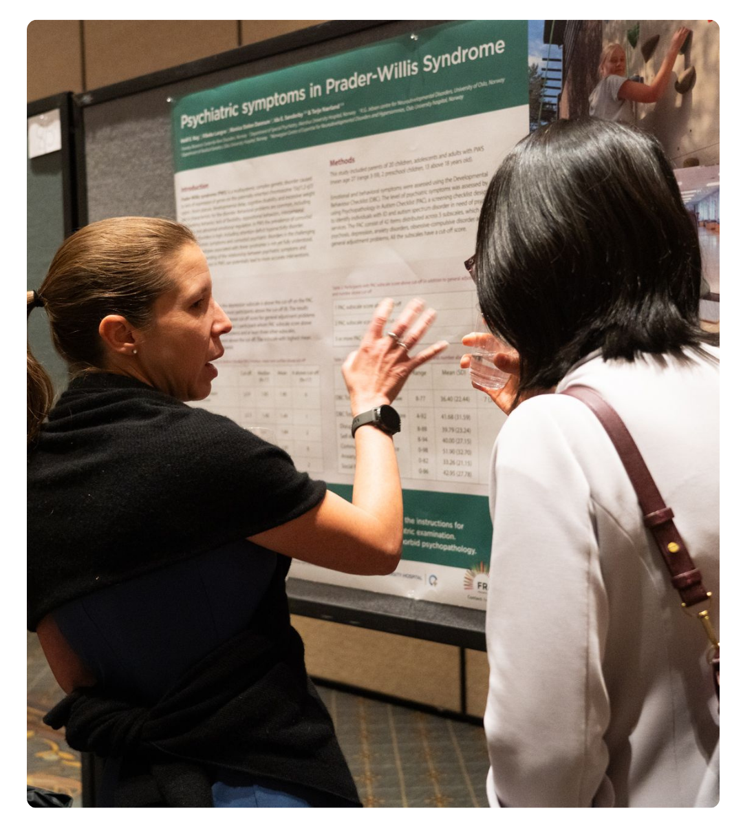 Two researchers discuss a scientific poster about psychiatric symptoms in Prader-Willi syndrome at a conference.